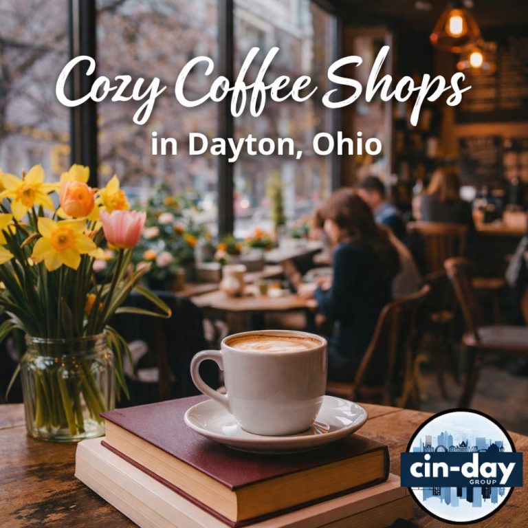 Latte in a white cup on a stack of books beside a jar of daffodils and tulips, with people relaxing in a warm, sunlit café behind it. Text reads "Cozy Coffee Shops in Dayton, Ohio."